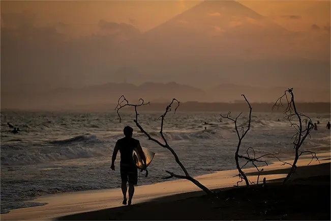 Surfer walking along a beach at sunset with mountains in the background