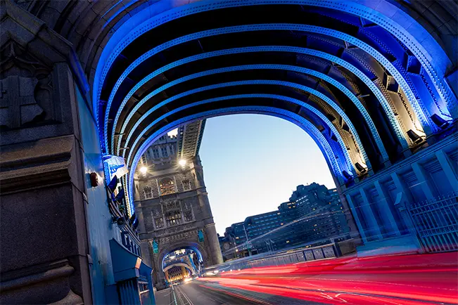 Light trails from passing vehicles beneath a blue illuminated bridge