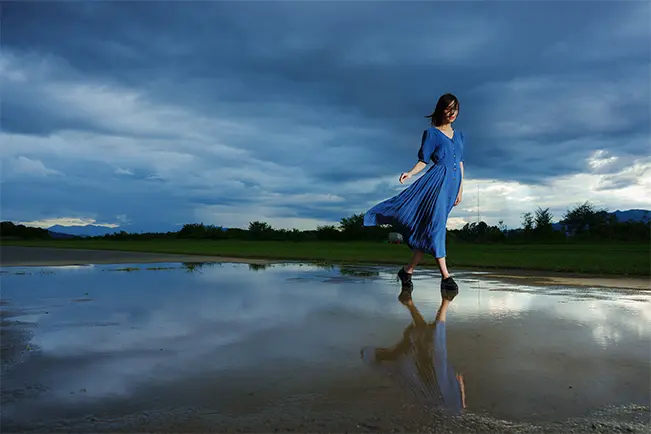 Woman in a blue dress walking across a reflective flooded field beneath a dramatic sky