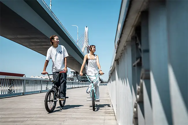 Man and woman riding bicycles along a waterfront promenade beneath an elevated bridge