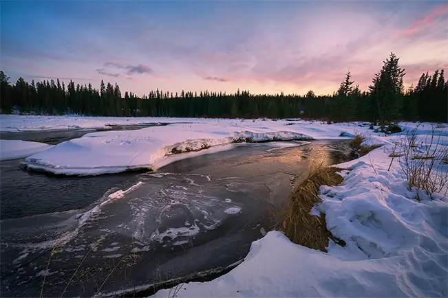 Snow-covered river flowing through a forest at sunset